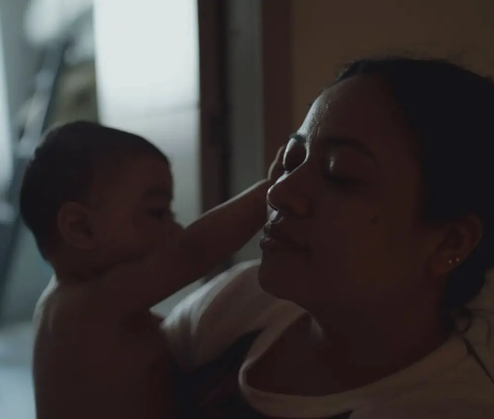 A mother closes her eyes in rest while her baby in her arms reaches out to her face.