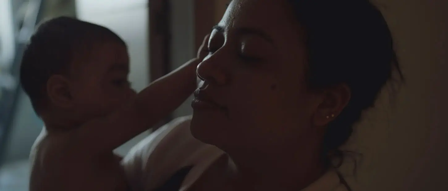 A mother closes her eyes in rest while her baby in her arms reaches out to her face.